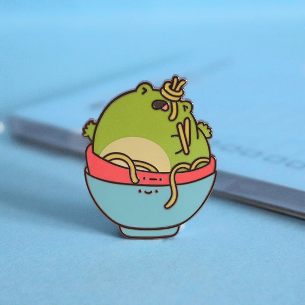 Frog in a bowl enamel pin on blue table with notepad