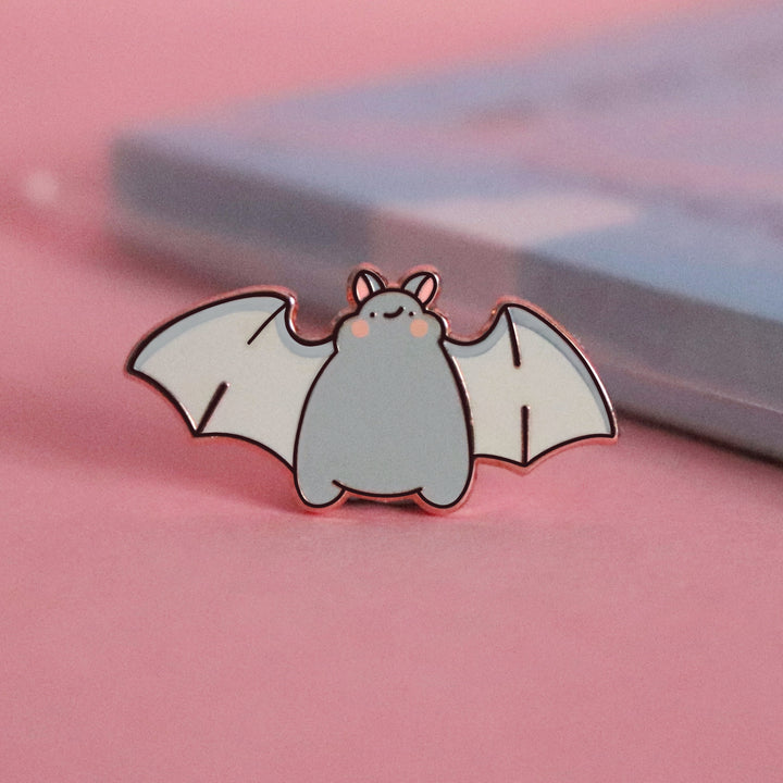 Bat smiling enamel pin on pink table with notepad