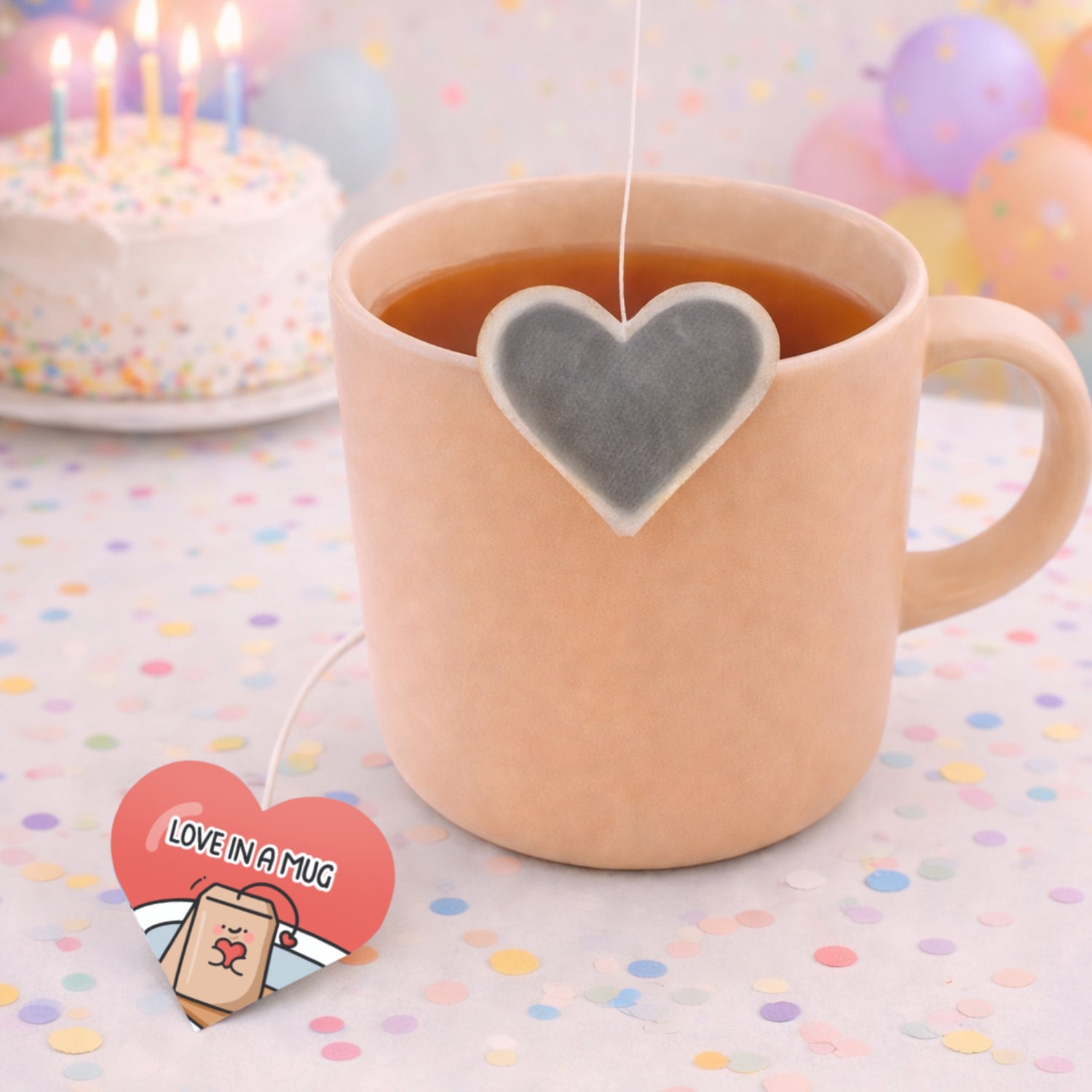 Pink mug with heart-shaped tea bag on a colorful polka dot tablecloth, with a birthday cake and balloons in the background.