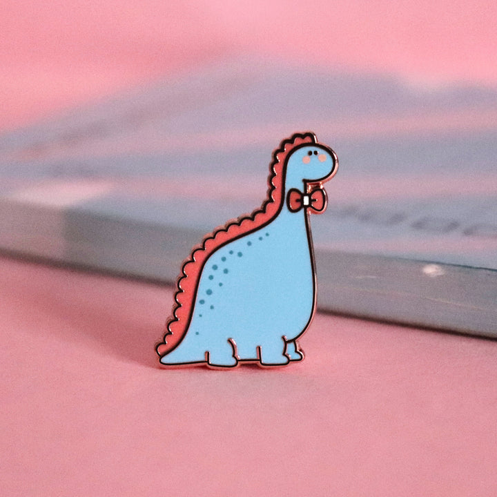 Blue dinosaur with bow tie enamel pin on pink table with notepad