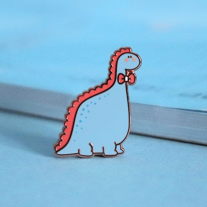 Blue dinosaur with bow tie enamel pin on blue table with notepad