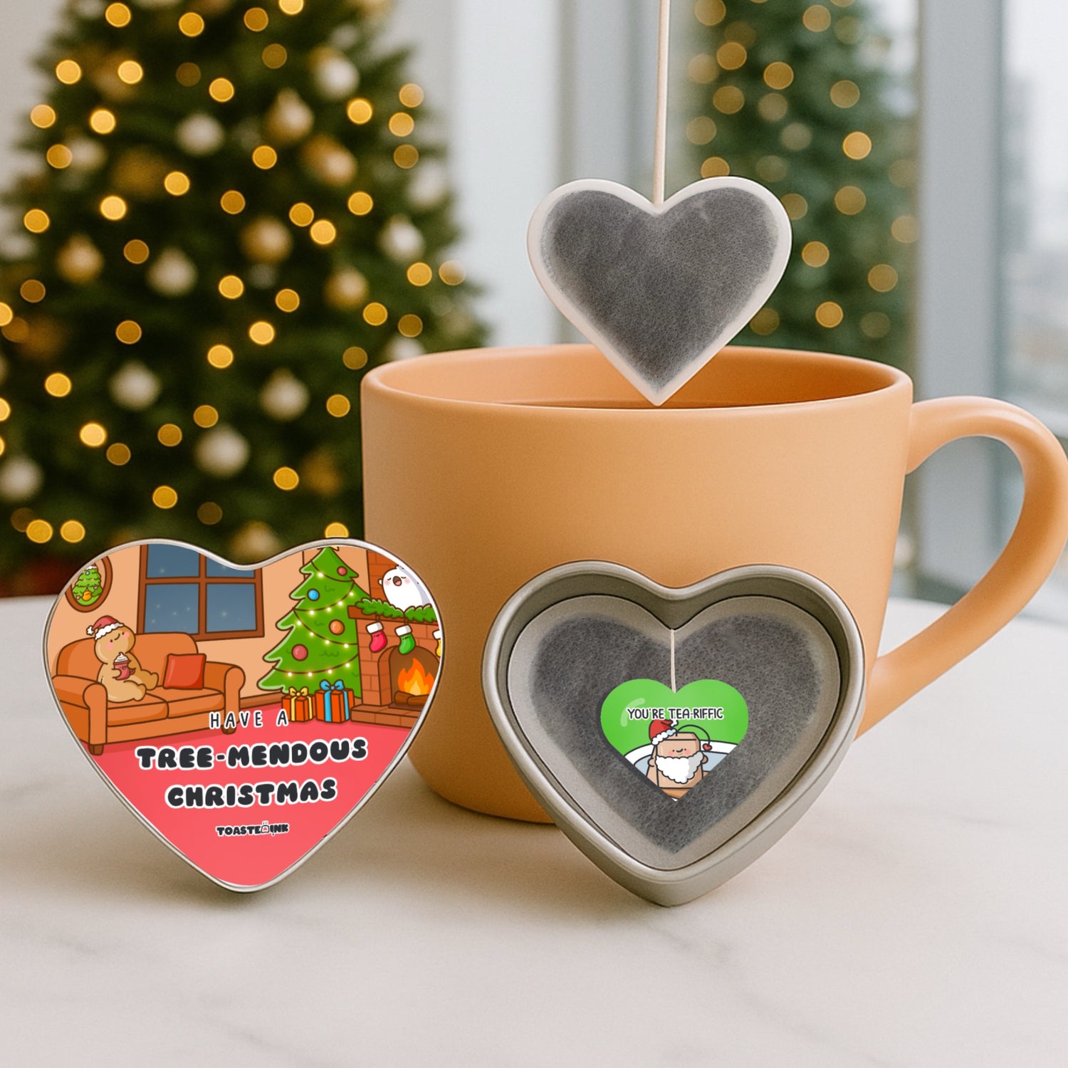 Heart-shaped tea bags with Christmas-themed designs in front of a mug and decorated Christmas tree.