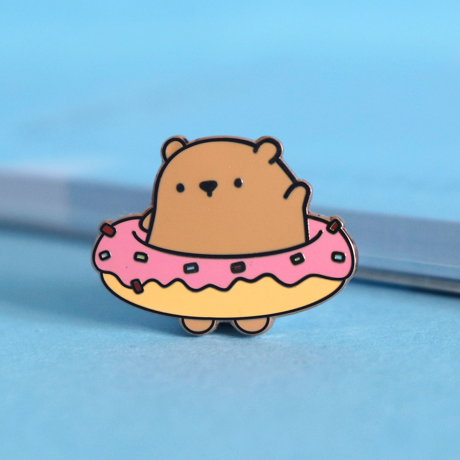 Bear wearing donut enamel pin on blue table with notepad