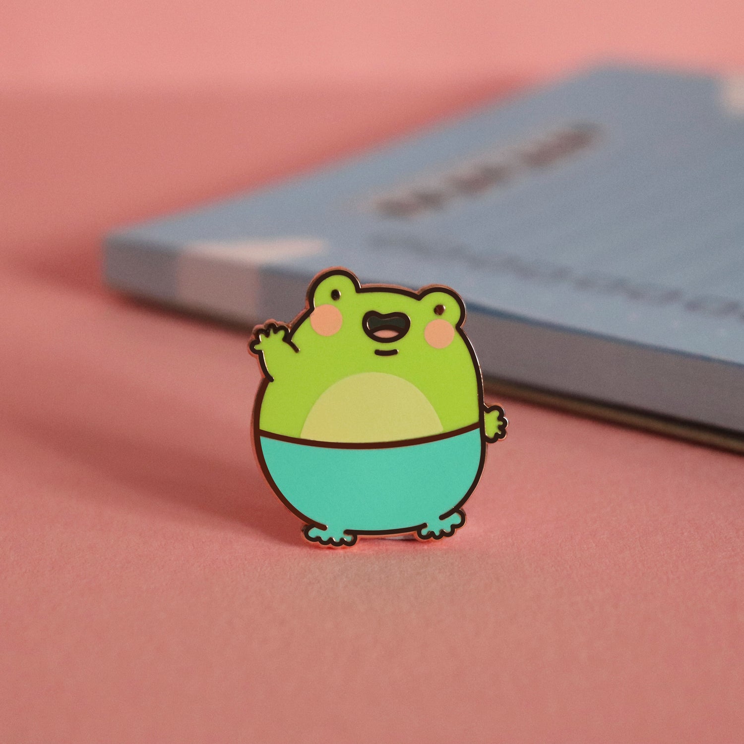 Frog enamel pin on pink table with notepad