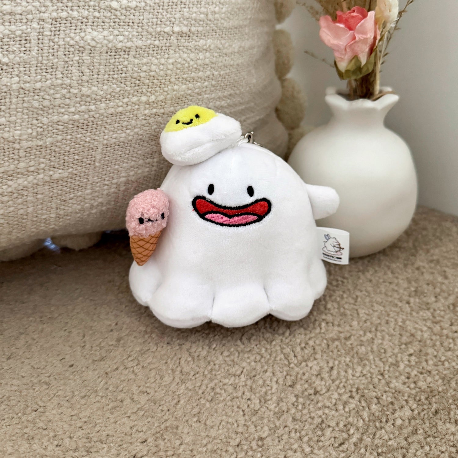 Cute ghost keychain plush toy sitting by a soft neutral cushion