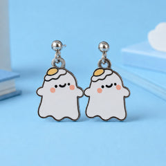 A pair of ghost-shaped stud earrings with a cute face, featuring a white body, yellow egg on its head, pink cheeks, displayed on a light blue background.