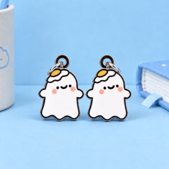 Two cute ghost-shaped earrings with white bodies, black outlines, pink cheeks, and yellow egg on his head details, displayed on a light blue background with a white cup and a blue notebook