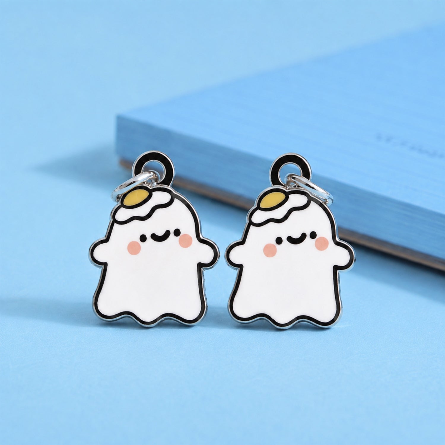 A pair of cute ghost-shaped earrings with dangle studs, featuring a white body, black outlines, pink cheeks, and a small yellow egg on his head on a light blue background.