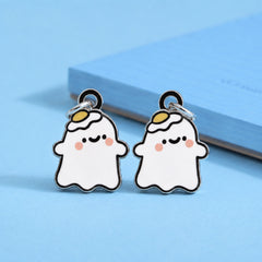 A pair of cute ghost-shaped earrings with dangle studs, featuring a white body, black outlines, pink cheeks, and a small yellow egg on his head on a light blue background.