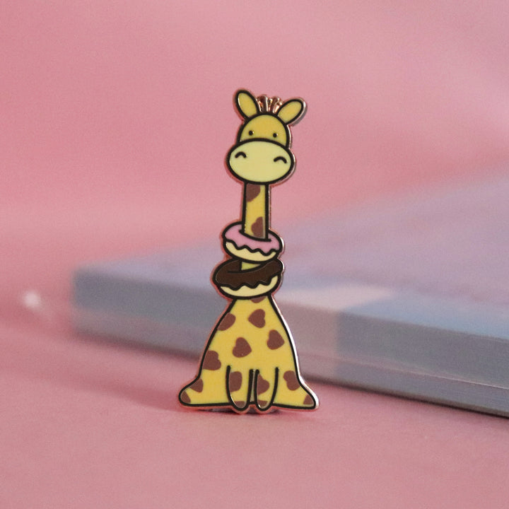 Giraffe with heart prints wearing donuts enamel pin on pink table with notepad