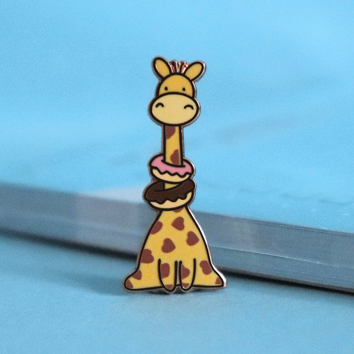 Giraffe with heart prints wearing donuts enamel pin on blue table with notepad