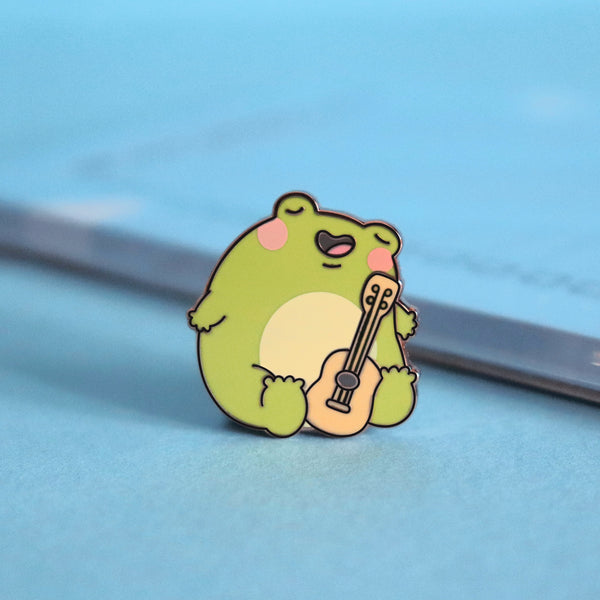 Frog with guitar enamel pin on blue table with notepad