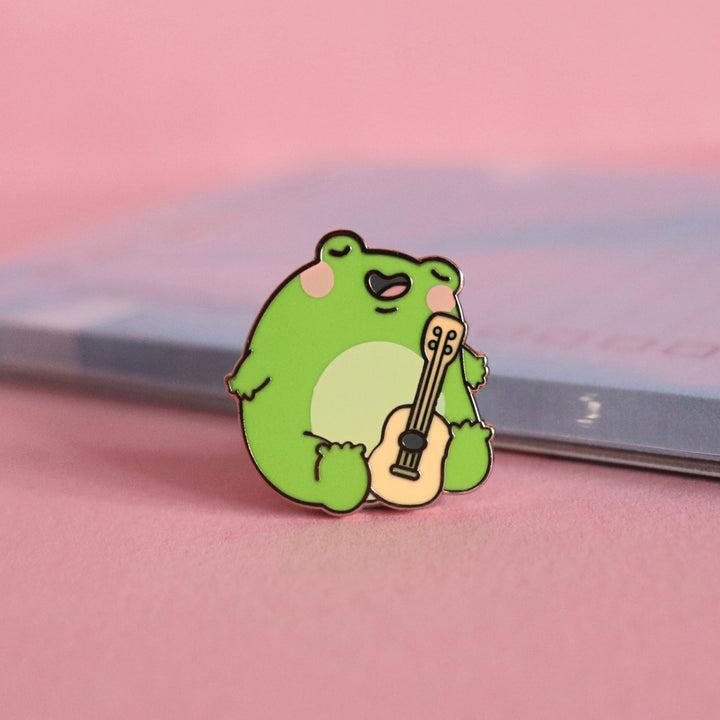 Frog with guitar enamel pin on pink table with notepad