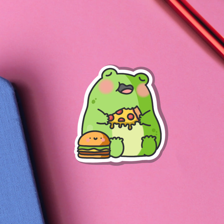Frog eating pizza vinyl sticker on pink table with notebook