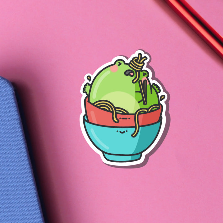 Frog stuck in noodles bowl vinyl sticker on pink table with notebook