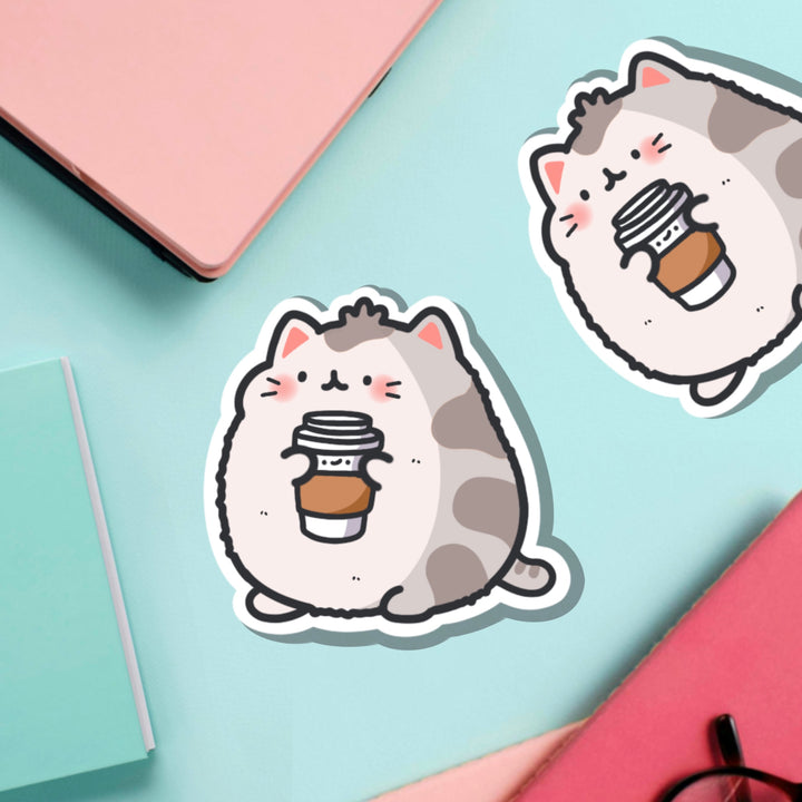 Cat holding coffee vinyl sticker on green table with notebooks