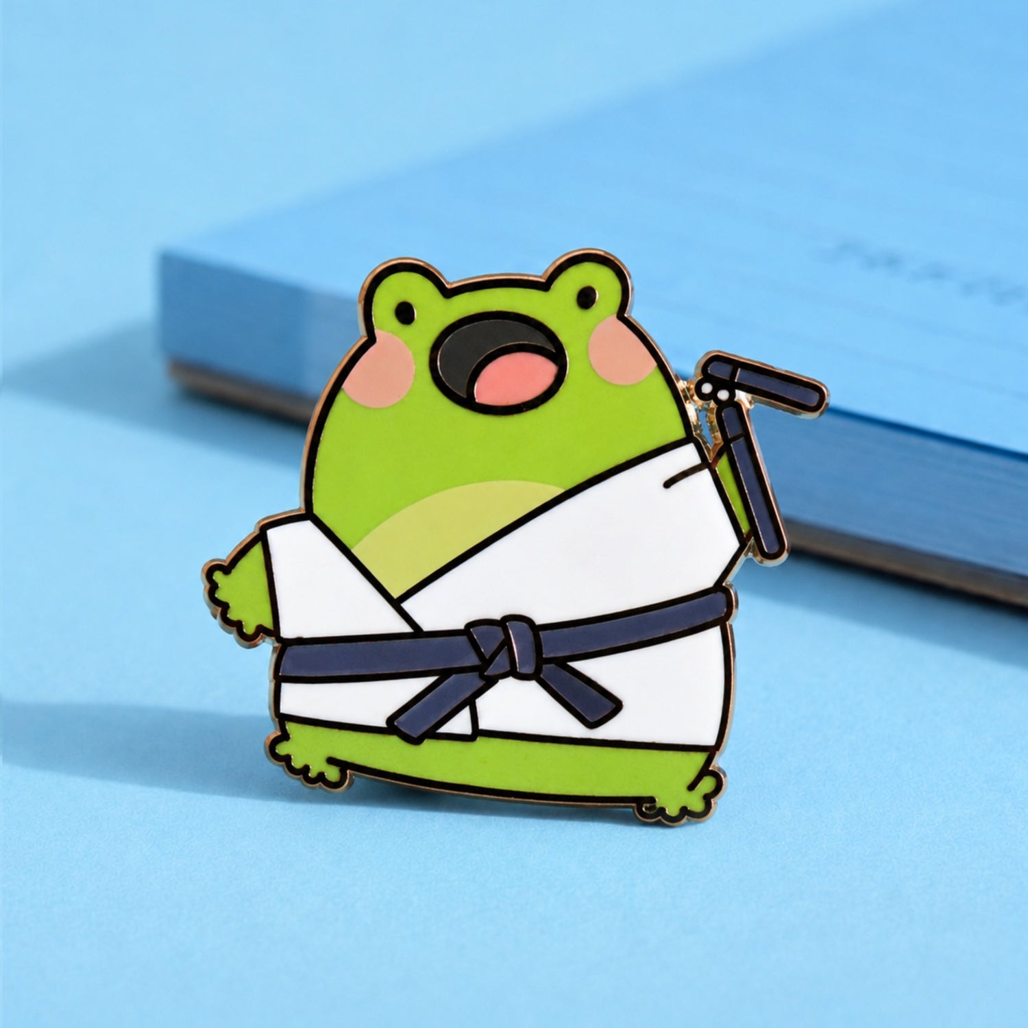 Cute karate frog enamel pin on blue table with notepad