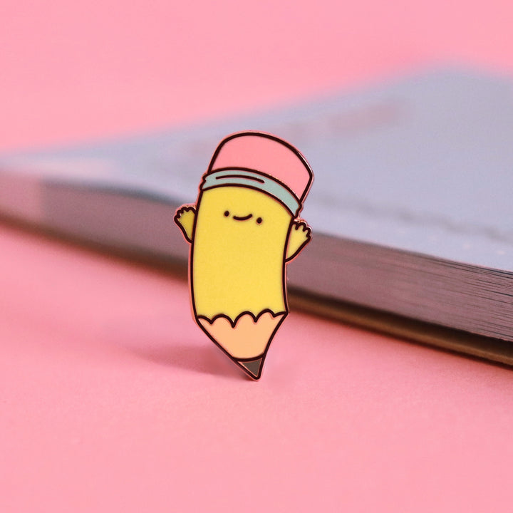 Pencil enamel pin on pink table with notebook