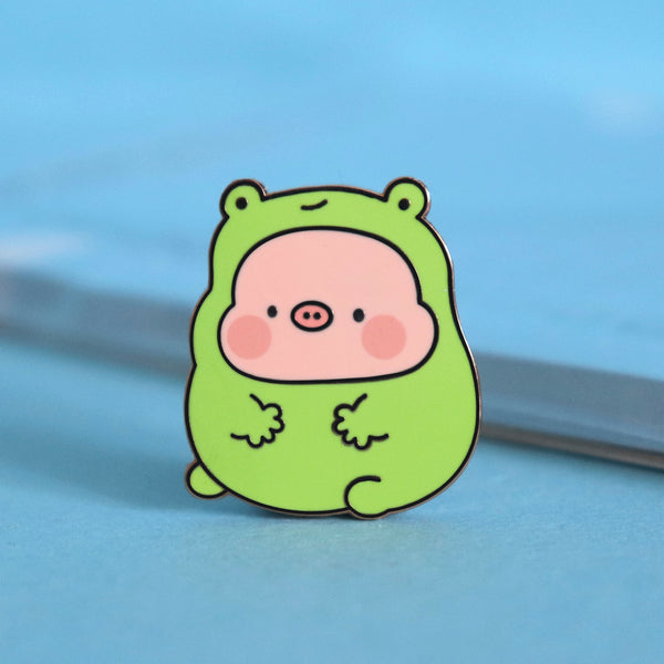 Pig in frog costume enamel pin on blue table with notepad