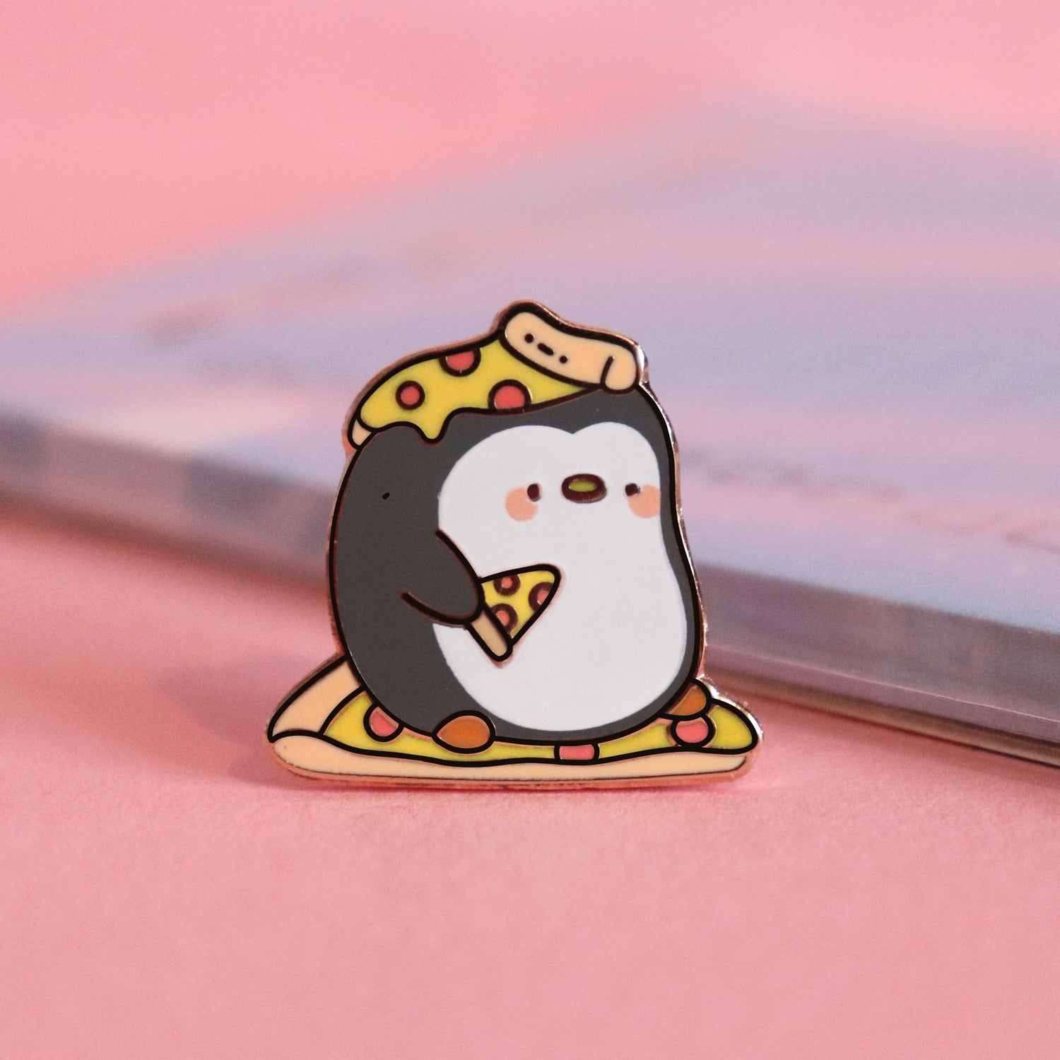 Penguin with pizza on his head enamel pin on pink table with notepad