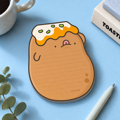 A cute potato-shaped notepad with lined writing space, placed on a light blue desk with a white mug, plant leaves, and a pen nearby.