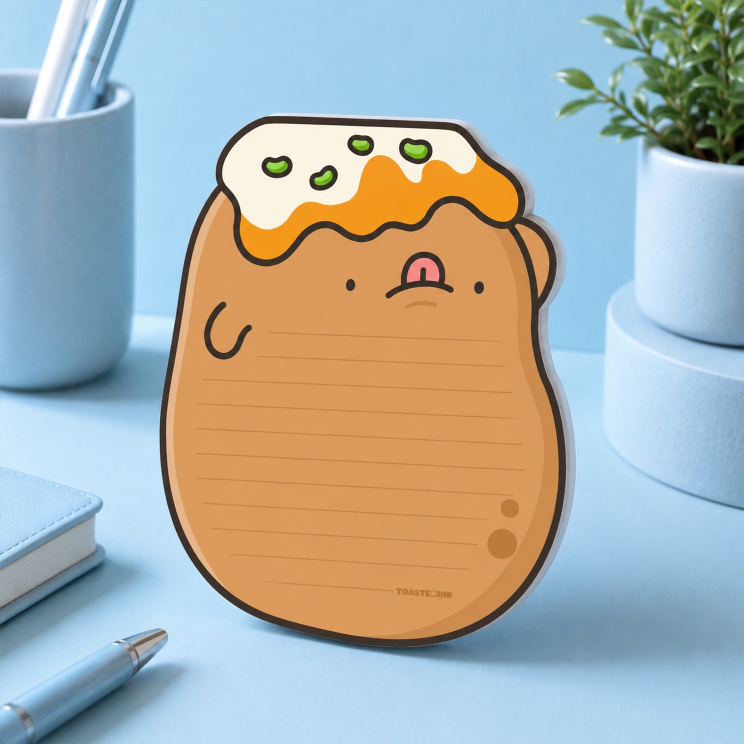 A cute potato-shaped lined notepad with a yellow topping details, standing on a light blue desk with office props in the background.