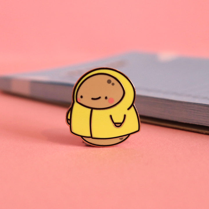 Potato in a raincoat enamel pin on pink table with notepad