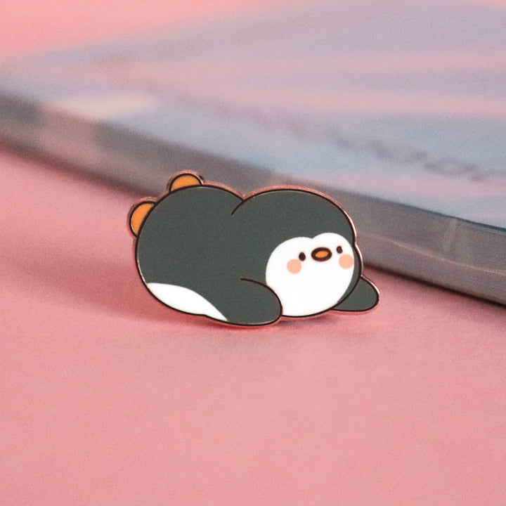 Penguin sliding on his belly enamel pin on pink table with notepad