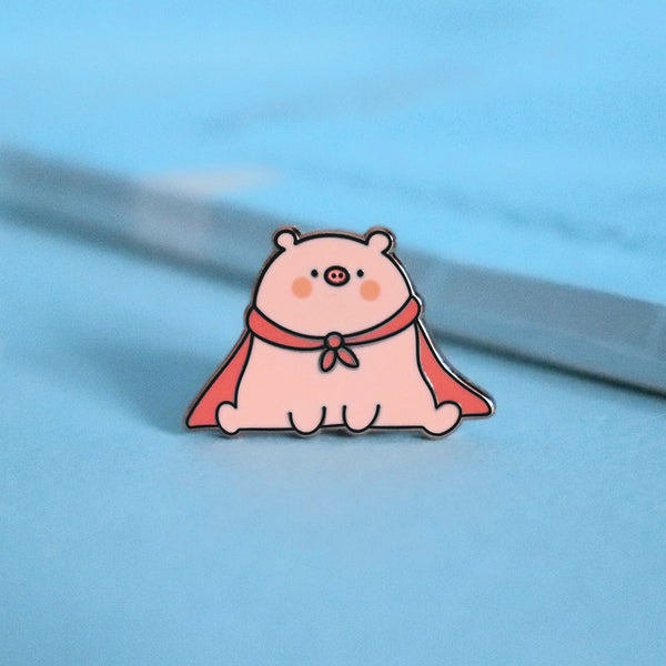 Super pig wearing red cape enamel pin on blue table