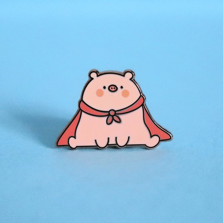 Super pig wearing red cape enamel pin on blue desk