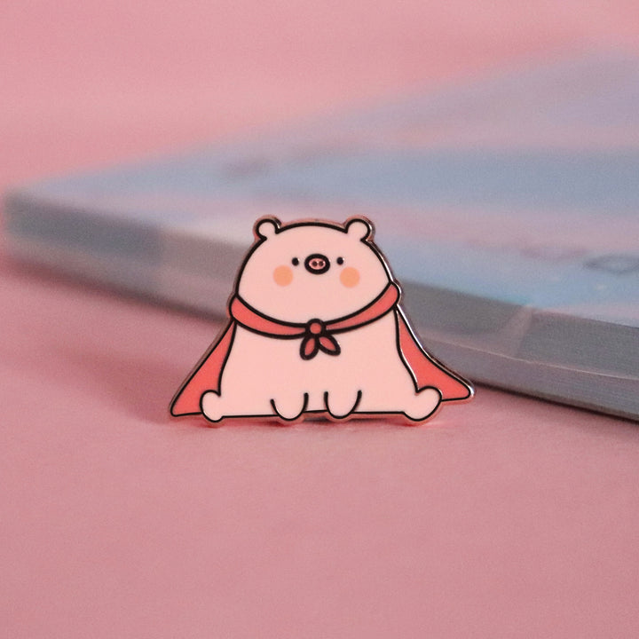 Super pig wearing red cape enamel pin on pink table with notepad