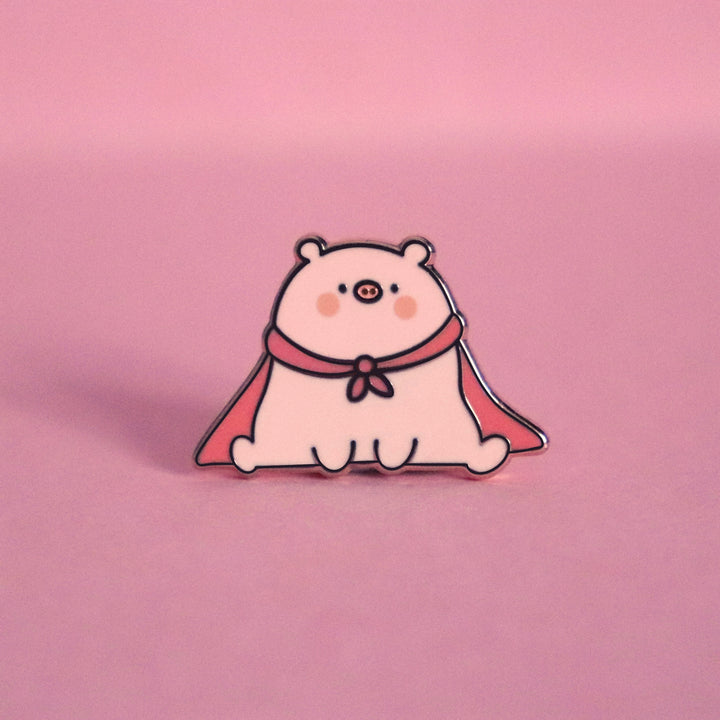 Super pig wearing red cape enamel pin on pink desk