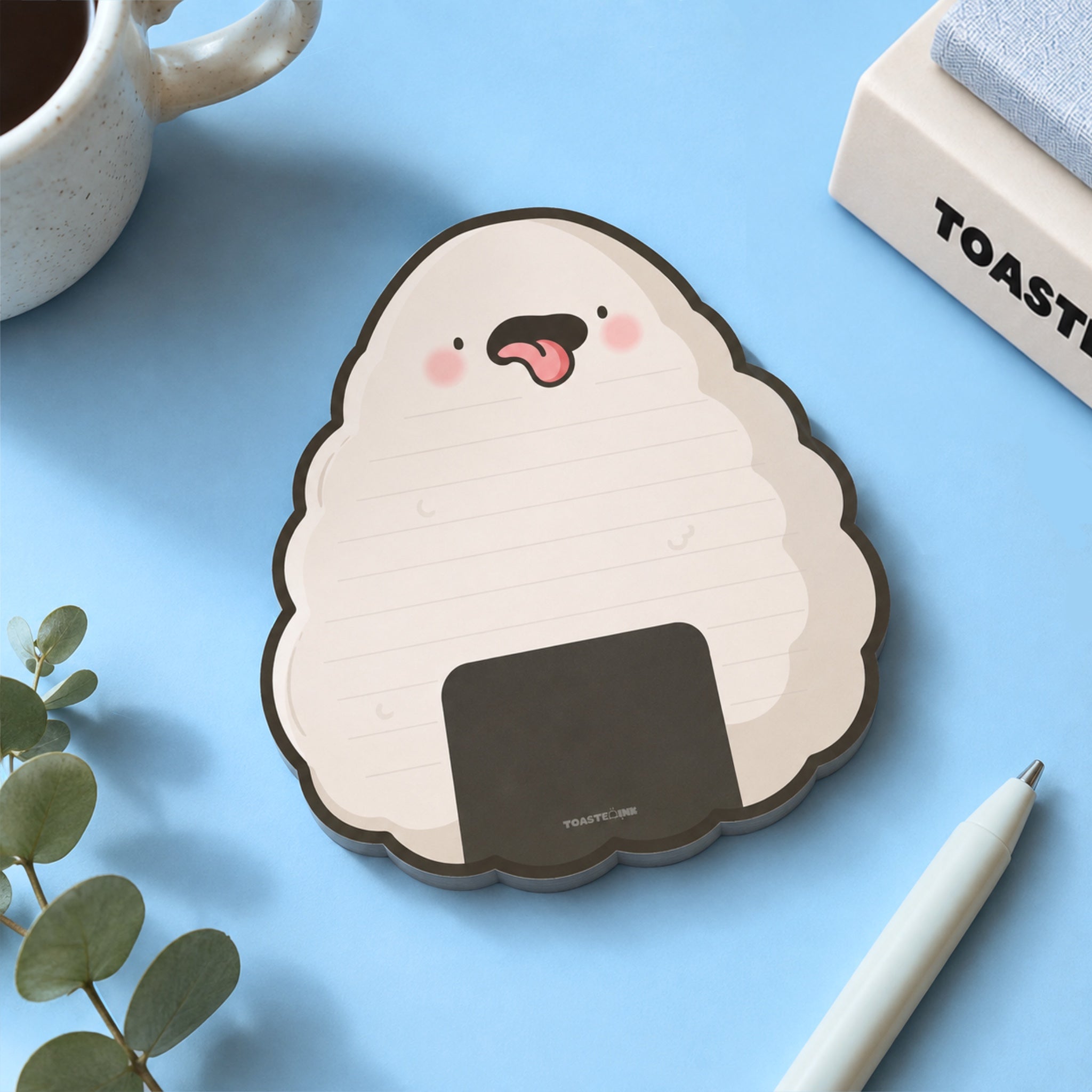 A cute onigiri-shaped notepad with a cheeky face, styled on a light blue desk with a white pen, a mug, and a small plant.
