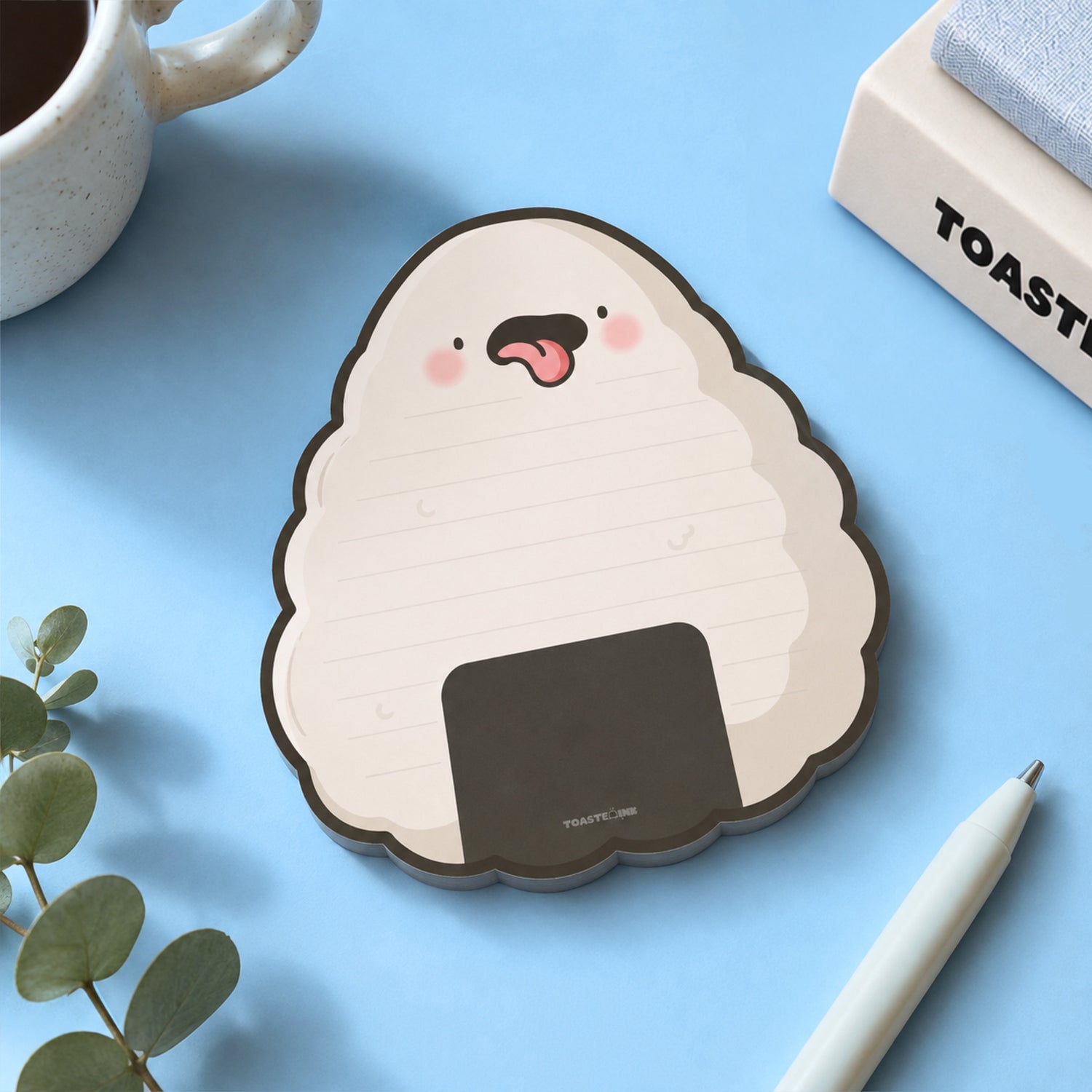 A cute onigiri-shaped notepad with a cheeky face, styled on a light blue desk with a white pen, a mug, and a small plant.