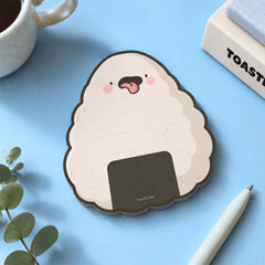 A cute onigiri-shaped notepad with a cheeky face, styled on a light blue desk with a white pen, a mug, and a small plant.