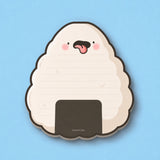 A cute onigiri-shaped notepad with a lined writing surface, pink cheeks, cheeky face, and a dark brown seaweed band, shown on a solid light blue background.