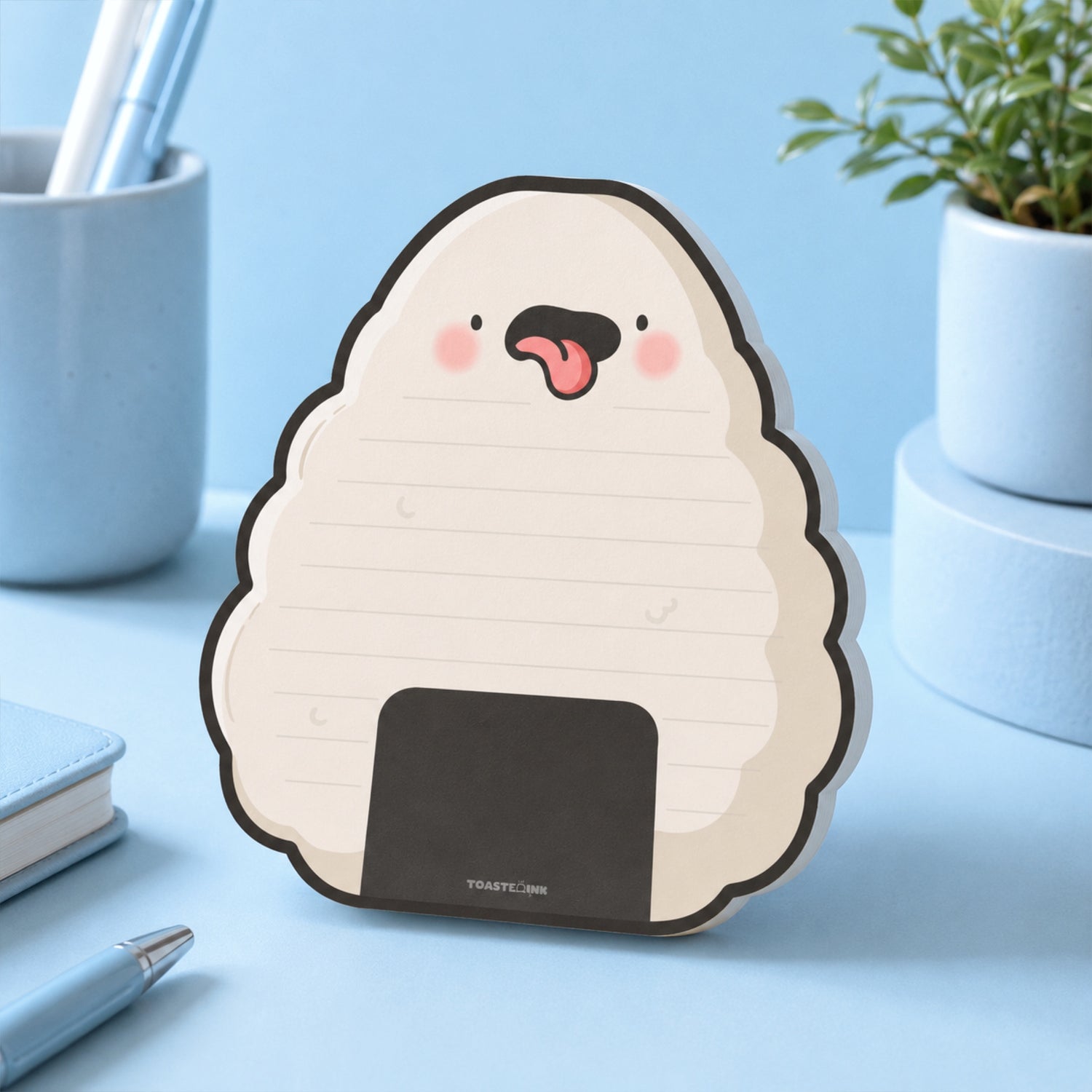 A cute onigiri-shaped desk notepad with a cartoon face, shown standing on a light blue desk with office props in the background.