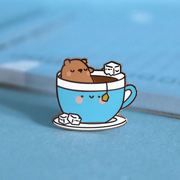 Bear sat in a cup of tea enamel pin on blue table with notepad