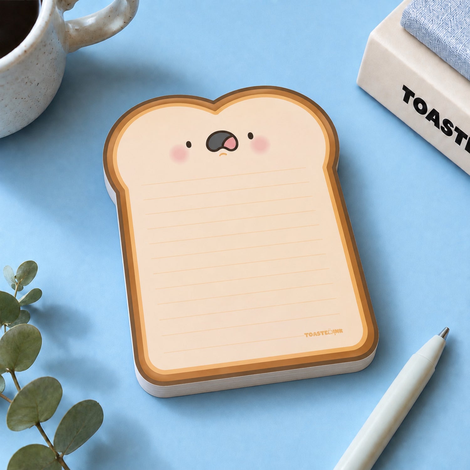 A cute toast-shaped notepad shown on a light blue background with a white pen, a mug, and a small plant