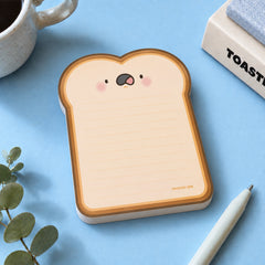 A cute toast-shaped notepad shown on a light blue background with a white pen, a mug, and a small plant