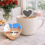 Heart-shaped 'I Love You' tea bag tag next to a cup of tea on a marble surface with flowers in the background.