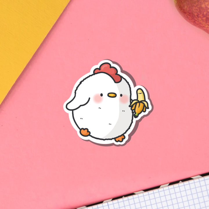 Chicken holding banana vinyl sticker on pink table with notebook