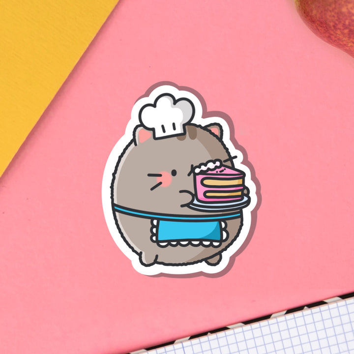 Cat holding cake vinyl sticker on pink table with notebook
