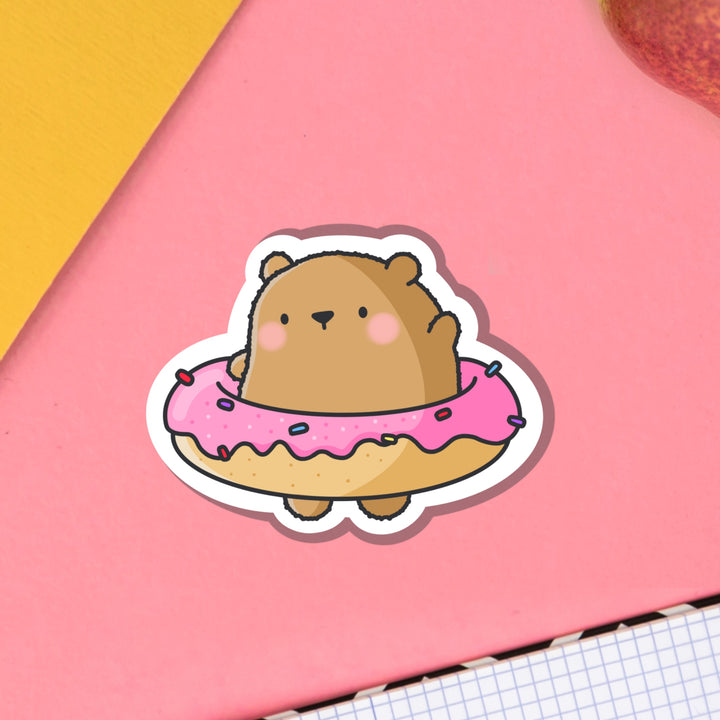 Bear wearing a donut vinyl sticker on pink table with notebook