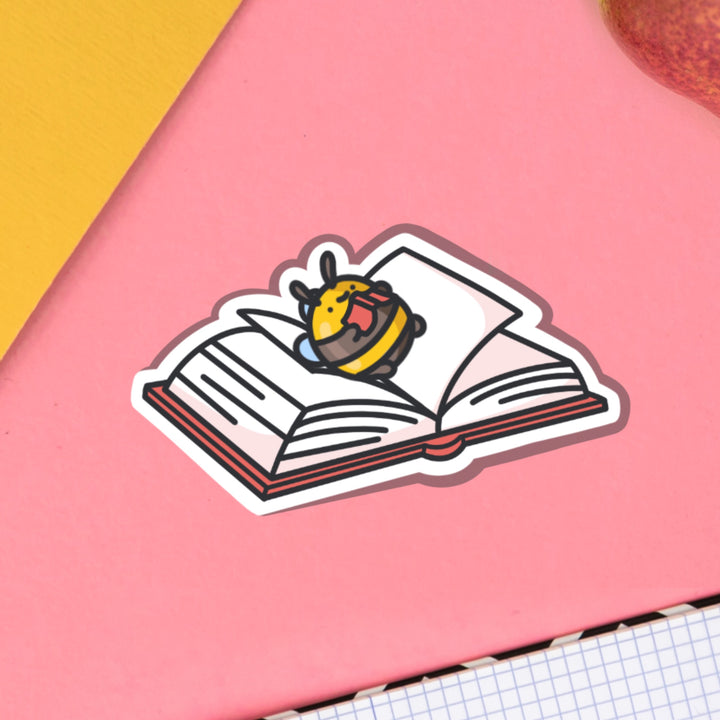 Bee lying on a book vinyl sticker on pink table with notebook