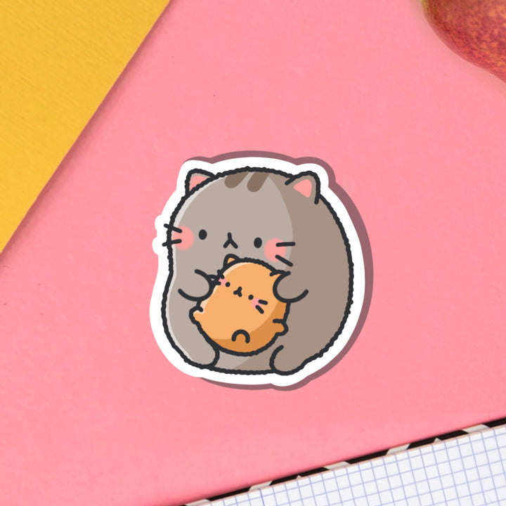 Cat holding baby kitty vinyl sticker on pink table with notebook