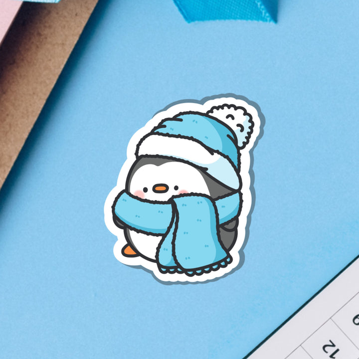 Penguin in scarf and wooly hat vinyl sticker on blue table